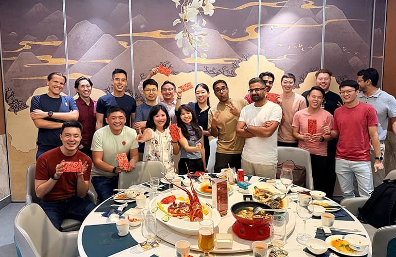 Group of colleagues gathered around a dining table sharing a meal, smiling and holding red envelopes, with dishes and drinks spread across the table.