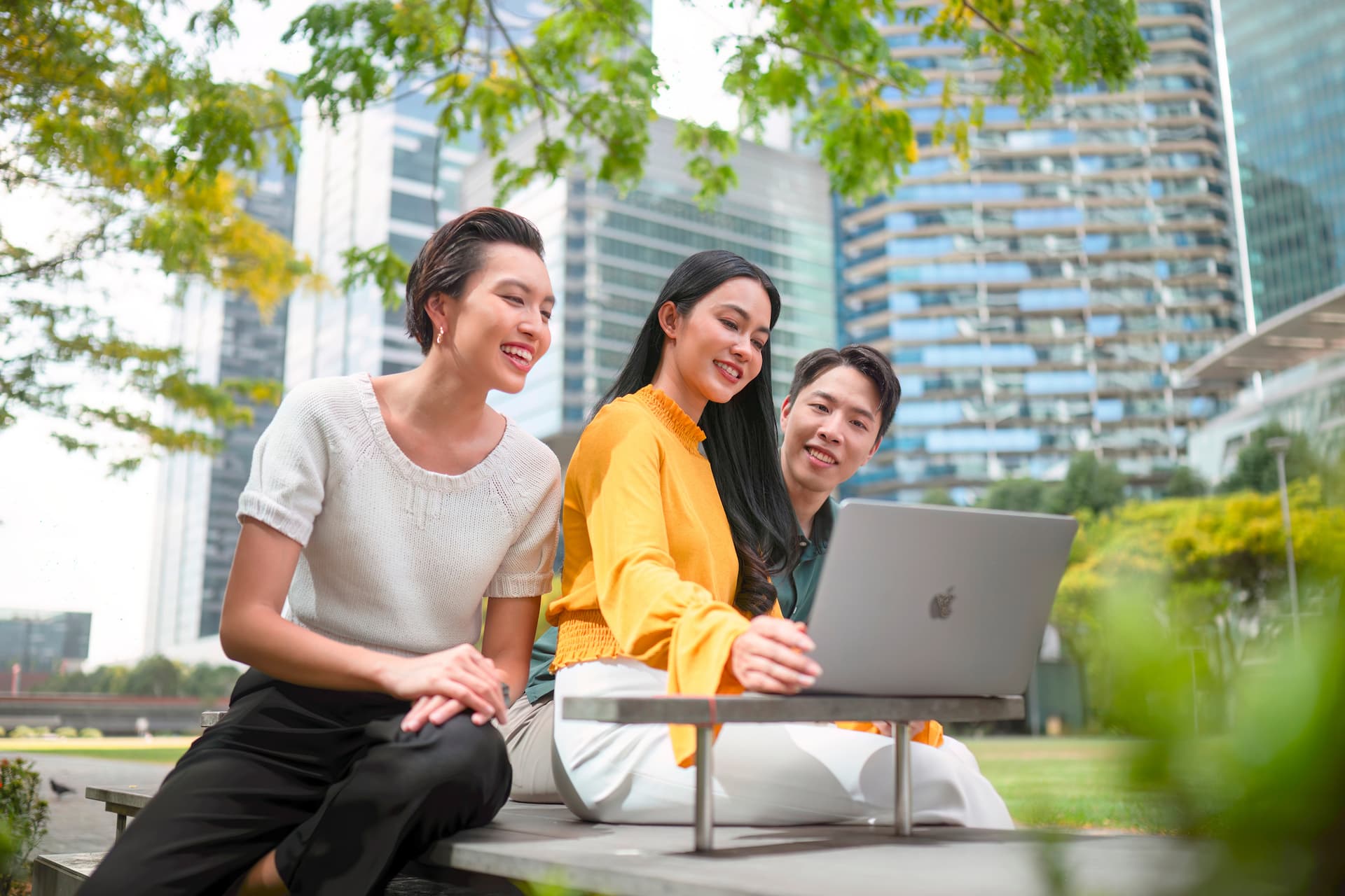 Three colleagues laughing over a laptop outdoors in Singapore.