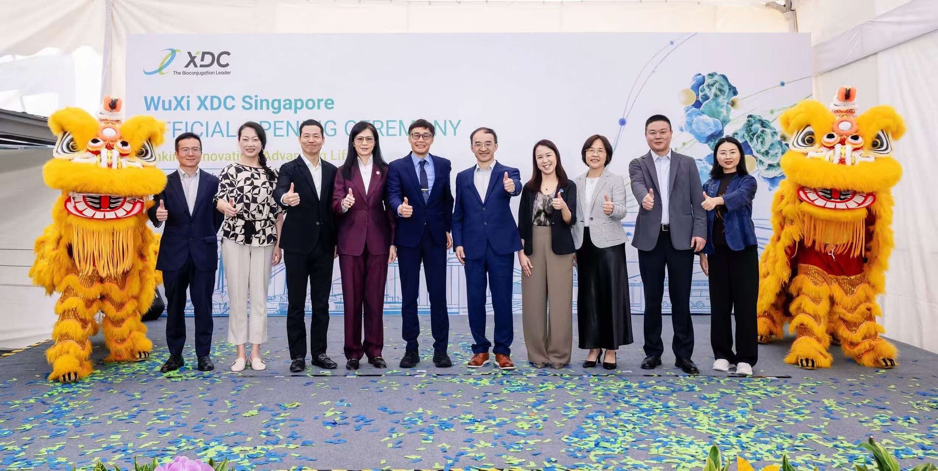 Corporate executives and two vibrant yellow lion dance performers pose together on a stage, giving thumbs-up gestures at the WuXi XDC Singapore Official Opening Ceremony.