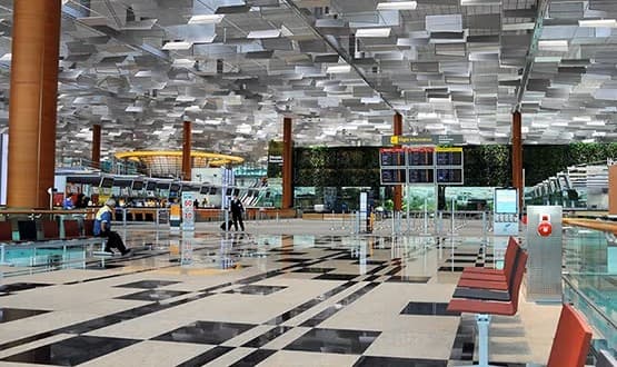 Spacious and modern airport terminal with geometric ceiling design, red seating, digital flight information boards, and a few travelers walking through the departure area.
