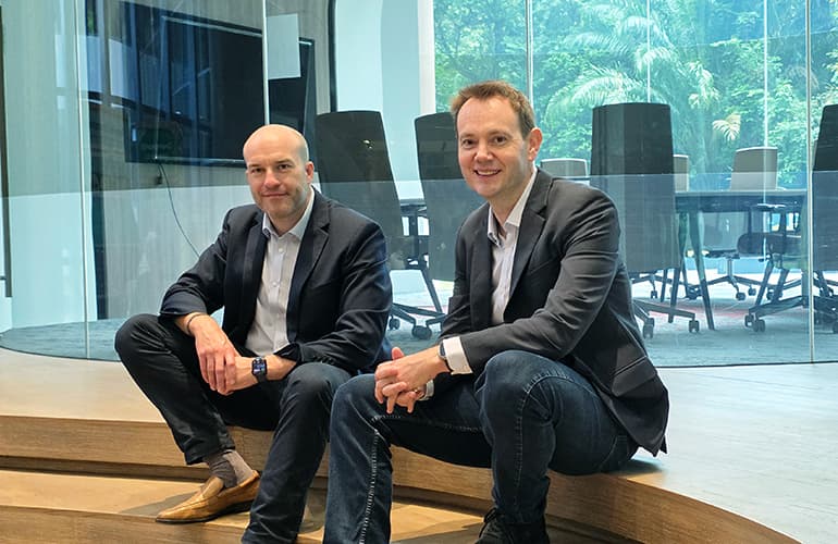 Two business executives seated in a modern office space with glass-walled meeting rooms and greenery visible in the background.