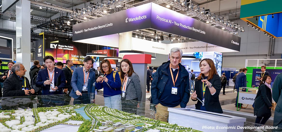 Visitors gather around a large scale model at a technology and industry exhibition, while exhibitors discuss innovation, manufacturing, and physical AI solutions.