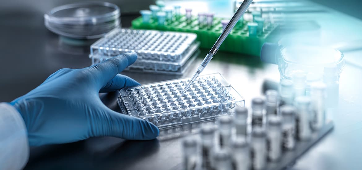 Scientist using a pipette to transfer liquid samples into a laboratory microplate, with vials and testing equipment arranged on a lab bench.