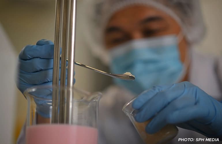 Scientist in protective gear carefully measuring powder into a beaker containing pink liquid during a laboratory experiment.