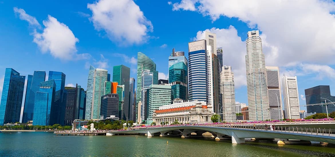 Singapore city skyline with high-rise buildings and a bridge over Marina Bay under a blue sky.
