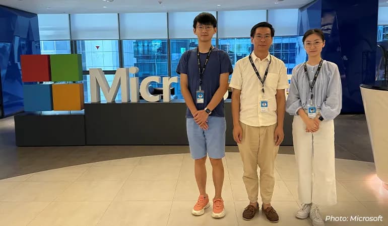 Three Microsoft staff members wearing ID badges stand in a modern office lobby in front of a large Microsoft logo, posing for a group photo with glass windows and city buildings in the background.
