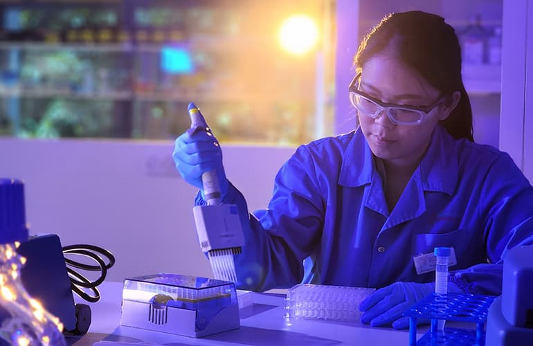 Scientist wearing protective eyewear and gloves using a pipette in a laboratory, surrounded by scientific equipment and sample trays under blue lighting.