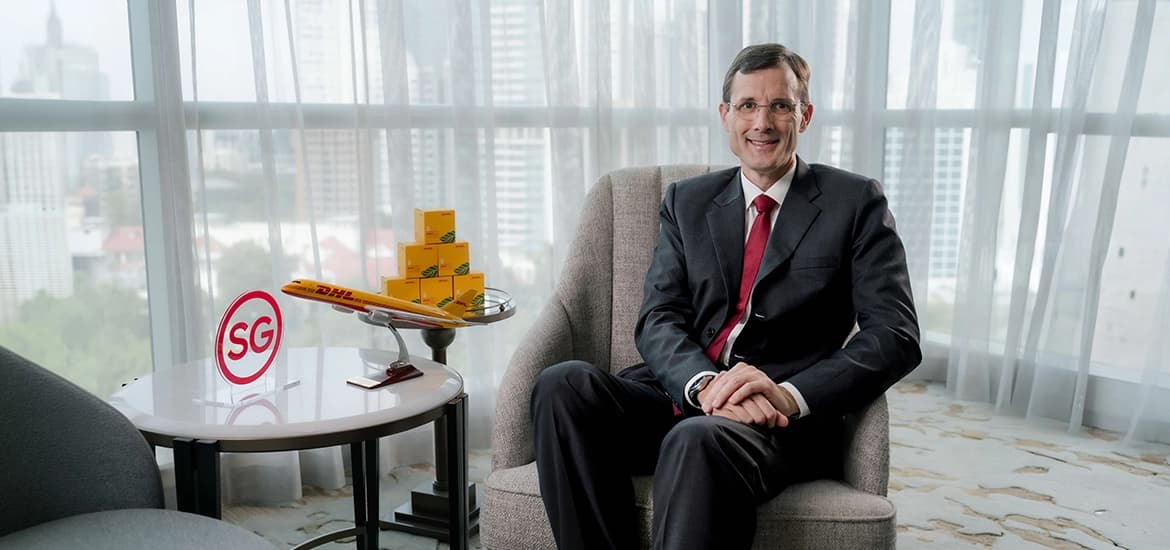 Tobias Meyer seated in a corporate setting, with a DHL model airplane and boxes on a side table alongside an “SG” sign, representing DHL’s Asia Pacific presence.