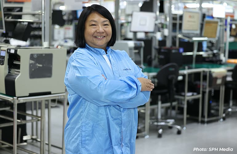 Woman in cleanroom attire standing with folded arms inside an electronics manufacturing facility, with production equipment and workstations in the background.