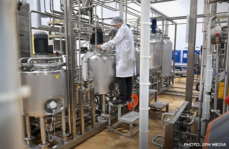Technician in a lab coat and hairnet inspecting large stainless-steel processing tanks inside a modern food and beverage manufacturing facility.