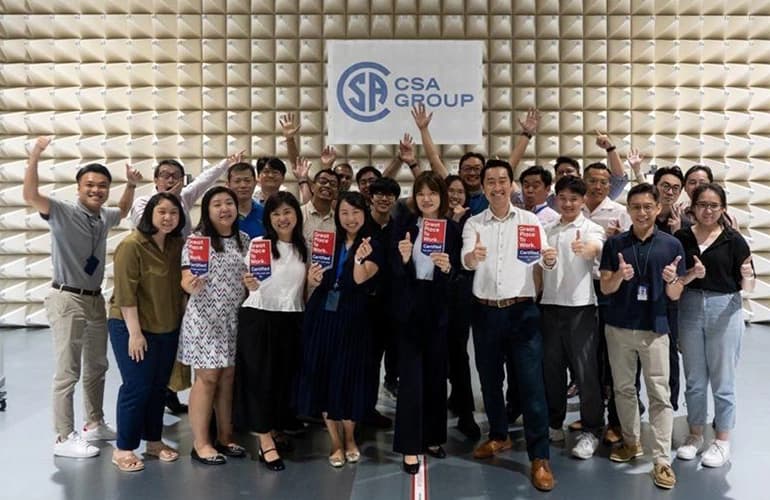 CSA Group team posing together indoors, smiling and giving thumbs up while holding booklets, with a CSA Group sign in the background.
