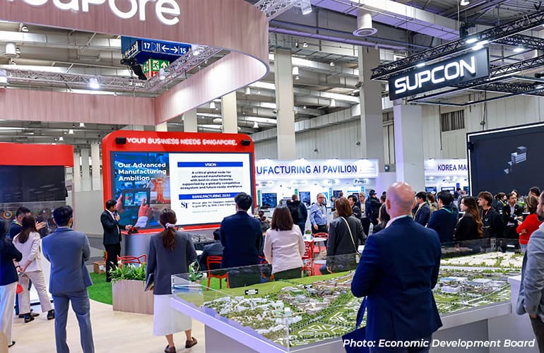 Visitors attending a Singapore exhibition booth focused on advanced manufacturing and AI, with a presentation screen, interactive displays, and industry pavilions in the background.