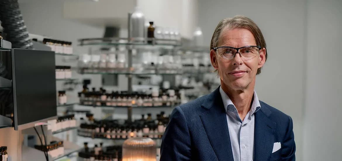 A man in business attire and safety glasses standing in a laboratory, with shelves of bottles and scientific equipment in the background.
