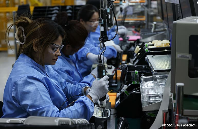 Workers wearing protective eyewear and cleanroom attire assembling electronic components on a production line in a manufacturing facility.