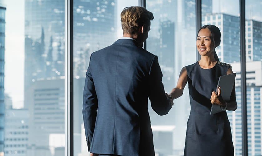 Two professionals shaking hands by a city skyline window.