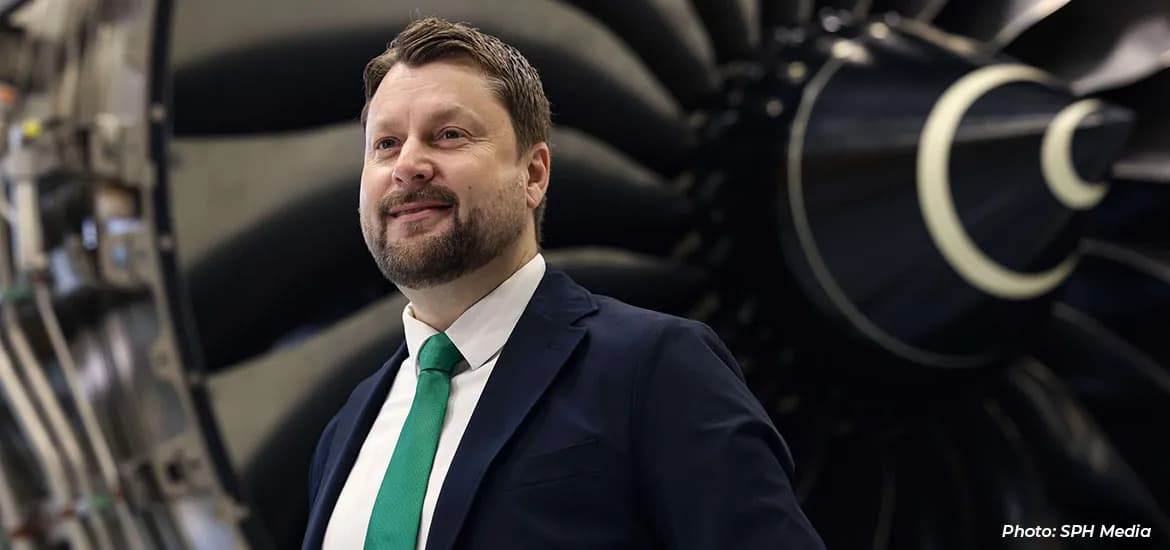 Man in a suit with a green tie standing in front of a large aircraft jet engine.