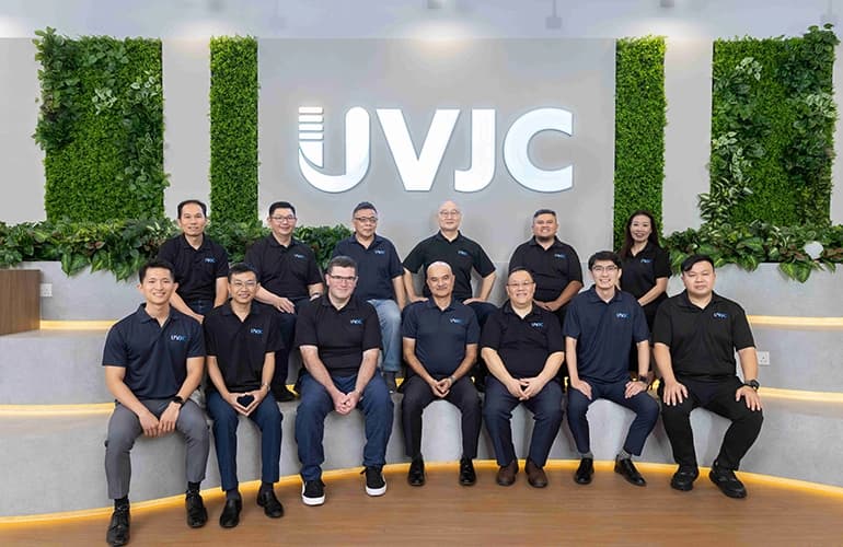 Group photo of the UVJC team seated and standing in an office space, with a large UVJC logo and greenery wall in the background.