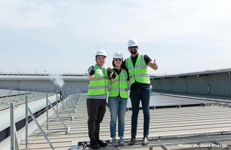 Three workers in safety vests and helmets standing on a rooftop solar installation.