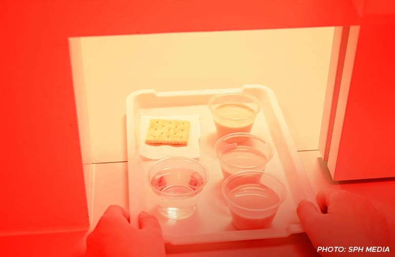 A person places a tray under red lighting containing four plastic cups with different liquids and a cracker on a napkin, prepared for a sensory or taste testing experiment.
