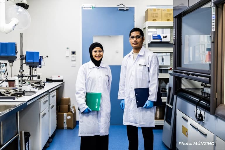 Two Münzing laboratory professionals in lab coats standing in a research lab, holding folders, with scientific equipment and workstations around them.