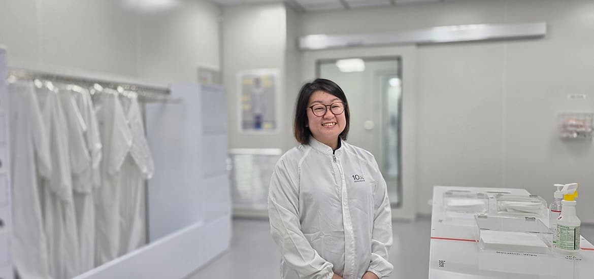 A technician in a cleanroom suit stands smiling inside a laboratory, with equipment and protective garments visible in the background.