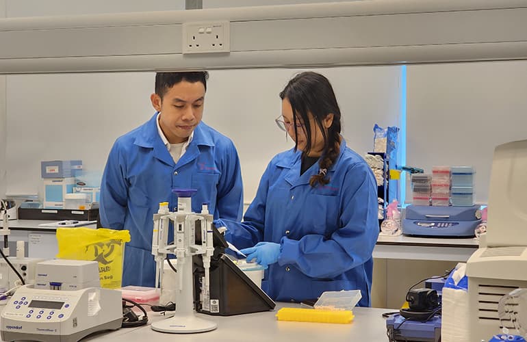 Two researchers in blue lab coats working together at a laboratory bench, operating scientific equipment and reviewing samples in a biomedical research facility.