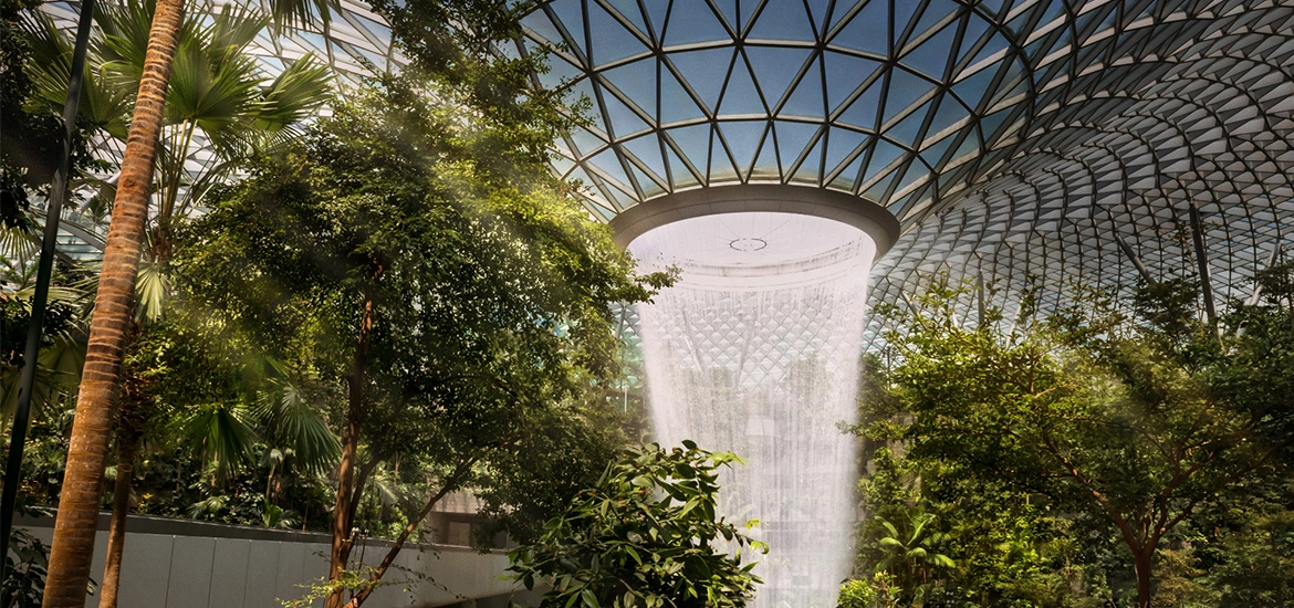 An indoor garden featuring a large central waterfall beneath a glass-domed roof, surrounded by lush greenery.