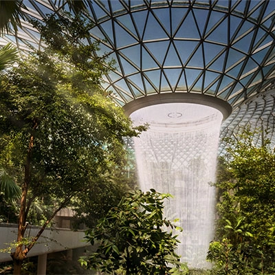 An indoor garden featuring a large central waterfall beneath a glass-domed roof, surrounded by lush greenery.