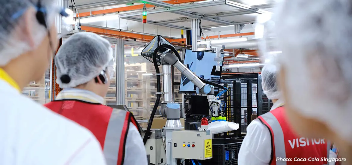 Workers in protective gear observe a robotic arm operating on a production line inside a manufacturing facility.