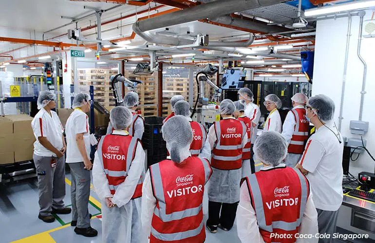 A group of visitors in protective gear and red vests observe automated machinery and robotic arms on a production line in a factory.