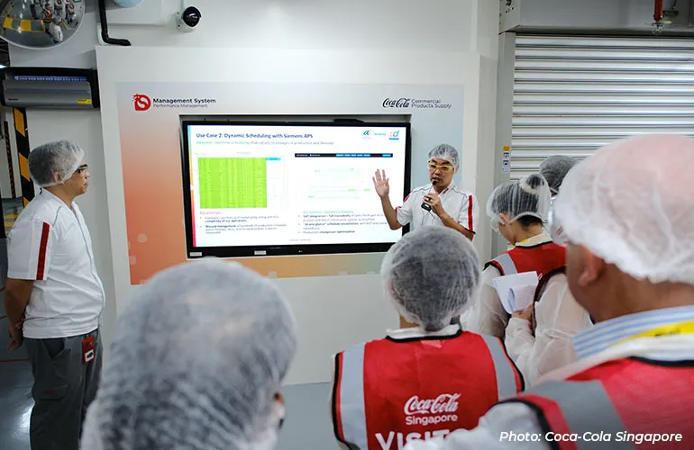 A presenter explains scheduling data on a screen to a group of visitors wearing protective gear inside a factory.