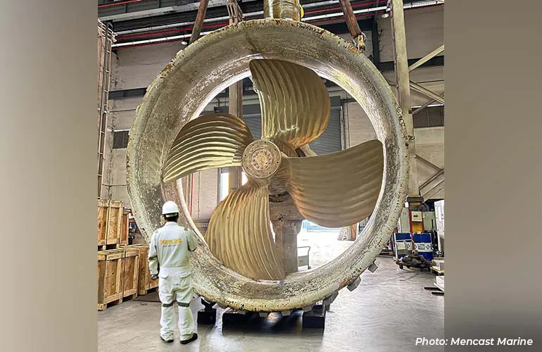 A worker stands beside a large ship propeller enclosed in a circular duct inside an industrial workshop.
