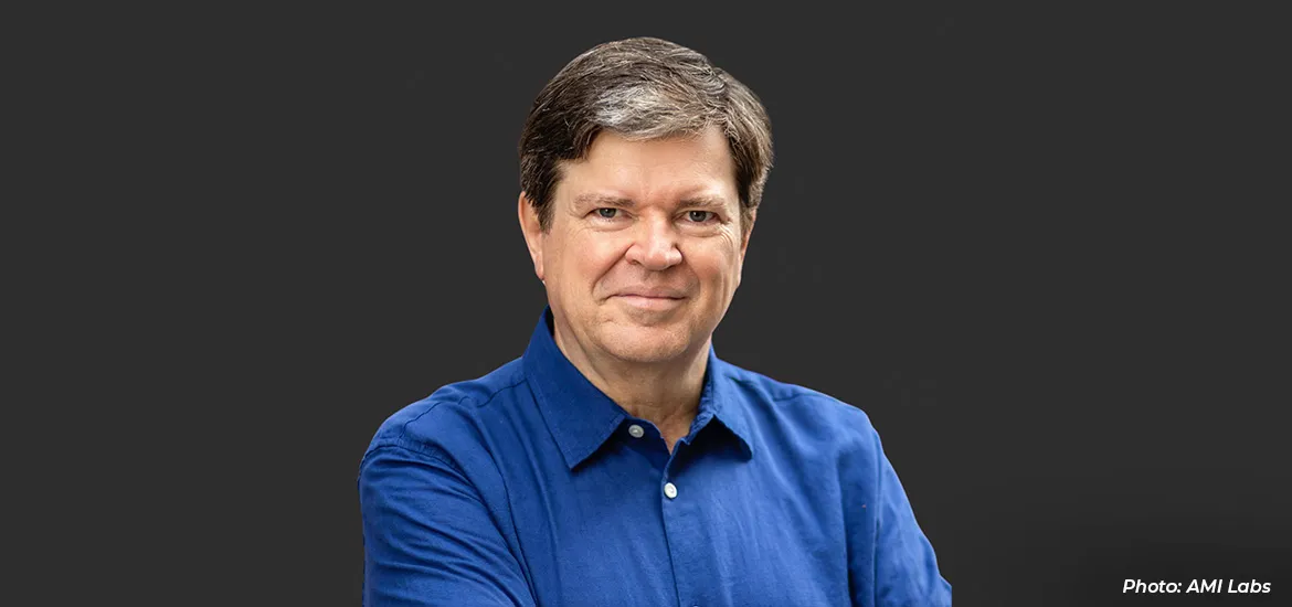 Portrait of a man in a blue shirt against a dark background, with arms crossed and a slight smile.