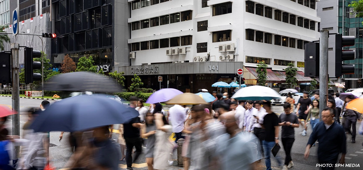 Crowds of people crossing a busy street in Singapore with colorful umbrellas during a rainy day, buildings and traffic lights in the background.