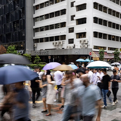 Crowds of people crossing a busy street in Singapore with colorful umbrellas during a rainy day, buildings and traffic lights in the background.