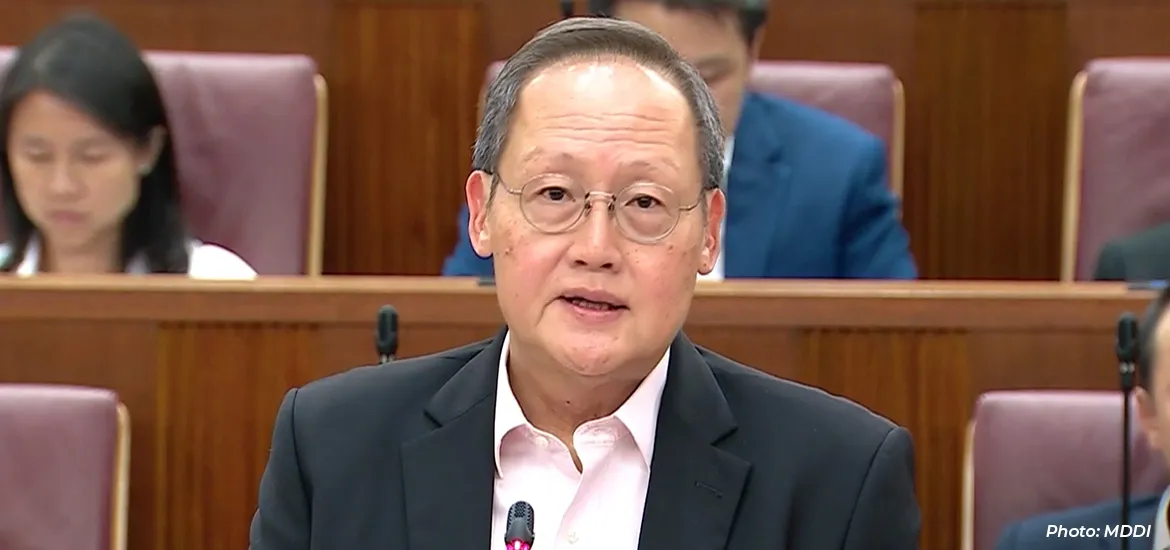 Man speaking at a podium during a parliamentary session, with seated members in the background.