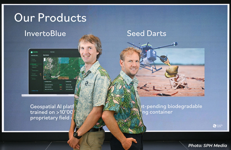 Two company representatives standing in front of a presentation display showcasing environmental technology products, including a geospatial AI platform for ecosystem monitoring and drone-based seed dispersal solutions for ecological restoration.