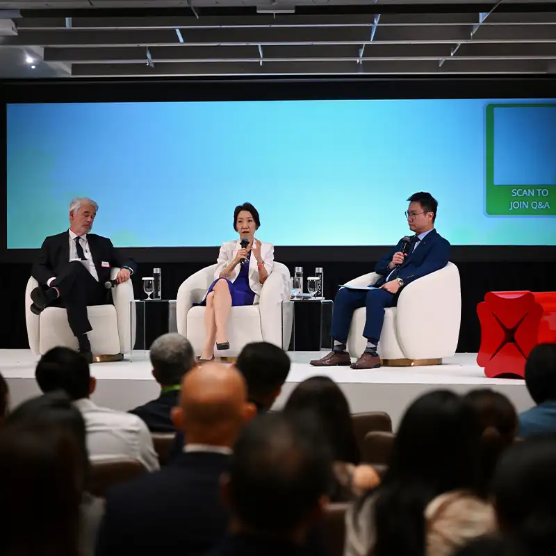 Panel discussion featuring three speakers seated on stage, with one speaker addressing the audience while holding a microphone during a business, sustainability, or industry conference.