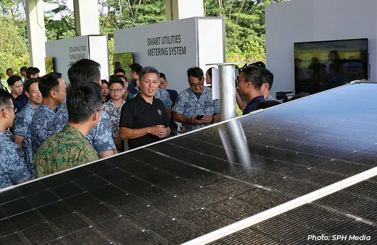 A group of people, including uniformed personnel, gathered around a solar panel installation while a presenter explains a smart utilities metering system display.