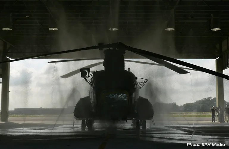 A helicopter silhouetted inside a hangar as water sprays over it, likely during a wash or maintenance process, with people standing nearby.