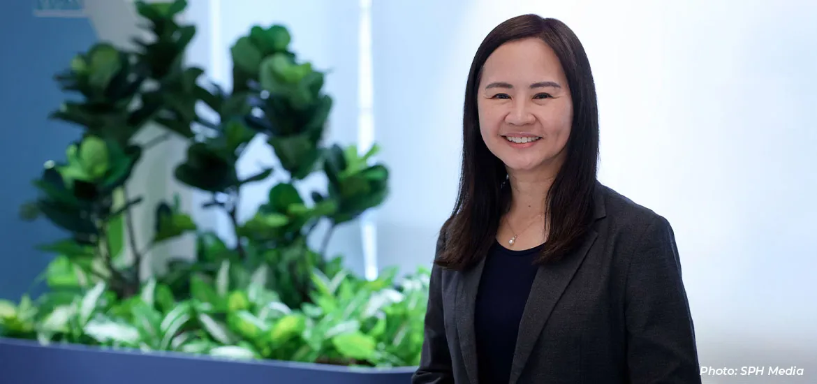 Smiling woman in a black blazer standing in a modern office setting with greenery in the background.