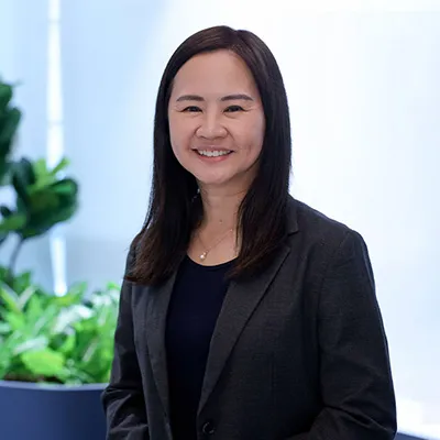 Smiling woman in a black blazer standing in a modern office setting with greenery in the background.