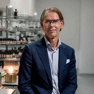 A man in business attire and safety glasses standing in a laboratory, with shelves of bottles and scientific equipment in the background.