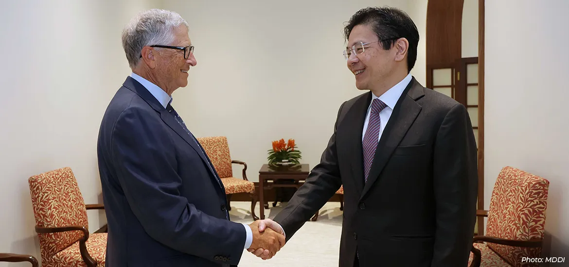 Two senior officials in business suits shake hands and smile during a formal meeting in an office setting, with upholstered chairs and a small table in the background.
