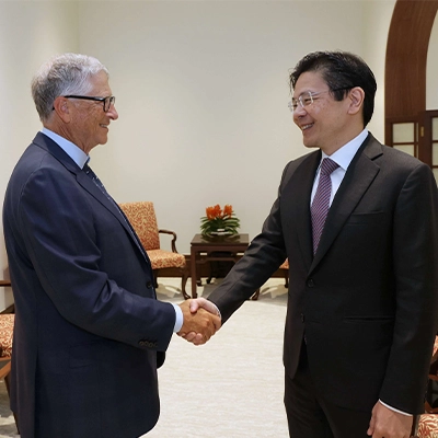 Two senior officials in business suits shake hands and smile during a formal meeting in an office setting, with upholstered chairs and a small table in the background.