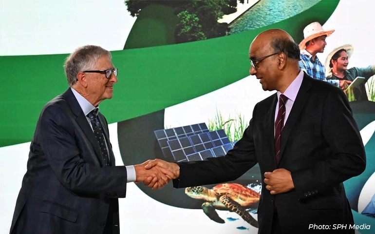 Two men in business suits shake hands and smile at a formal event in Singapore, with a backdrop featuring sustainability-themed imagery such as solar panels, agriculture, and nature.