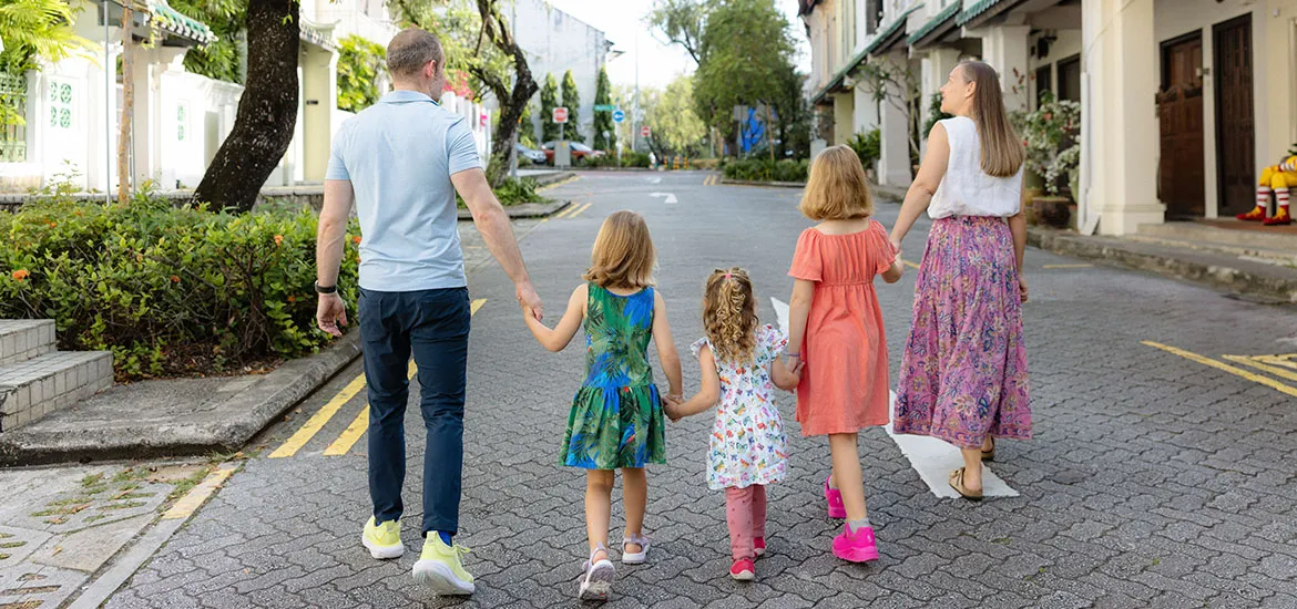 Family with three children walking hand in hand down a quiet, tree-lined street surrounded by greenery and traditional-style buildings.