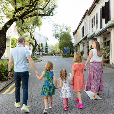 Family with three children walking hand in hand down a quiet, tree-lined street surrounded by greenery and traditional-style buildings.