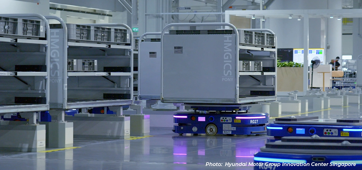 Autonomous mobile robots transporting storage racks inside a high-tech manufacturing facility at Hyundai Motor Group Innovation Center Singapore.