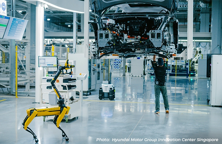Worker inspecting the underside of a car on an assembly line in a smart factory, with a robotic dog and digital control systems nearby at Hyundai Motor Group Innovation Center Singapore.